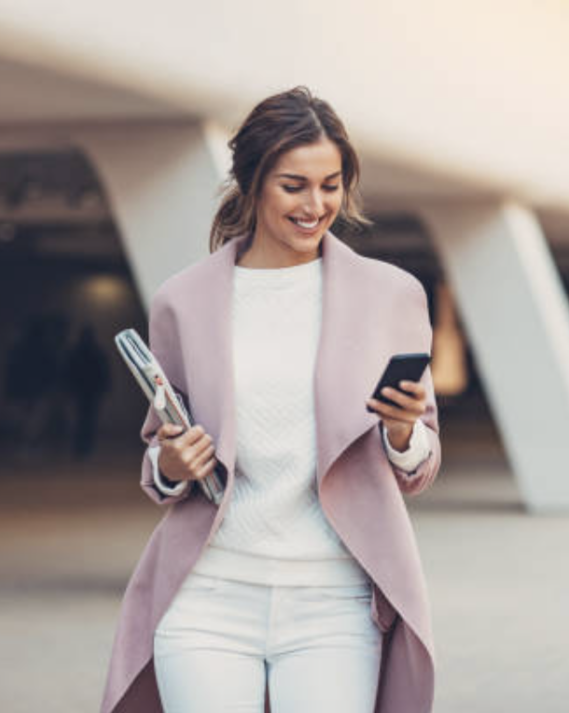 Vrouw in een roze jas kijkt op haar smartphone terwijl ze een flesje vasthoudt.